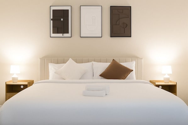 Una cama bien hecha con ropa blanca y dos almohadas, una mesita de noche con lámpara, y tres cuadros enmarcados en la pared.