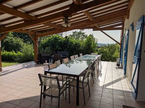 Une terrasse extérieure avec une longue table et des chaises, entourée d’une pergola en bois et offrant une vue sur la verdure et les collines.