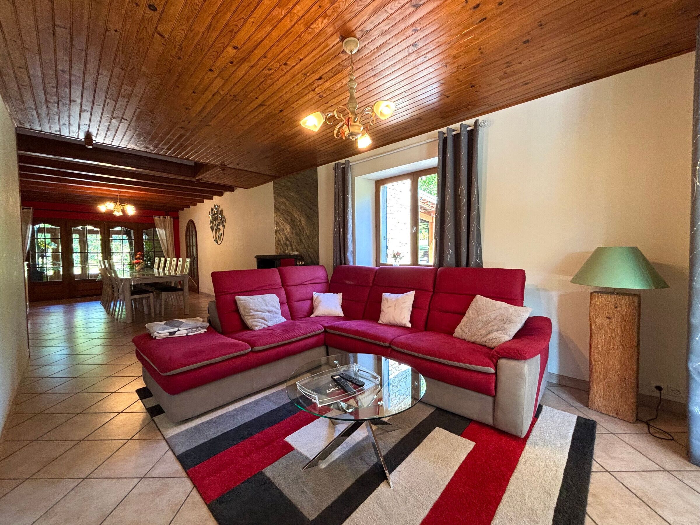 Una sala de estar acogedora con un sofá seccional rojo, una mesa de centro de vidrio y un tapete a rayas. El techo es de madera y hay una ventana con cortinas.