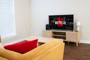Een woonkamer met een televisie waarop het Netflix-logo te zien is, een houten kast, een gele bank en een rood kussen.