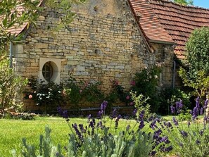 Un bâtiment en pierre, surmonté d’un toit recouvert de tuiles rouges, entouré d’un jardin bien entretenu où poussent de la lavande et d’autres plantes à fleurs.