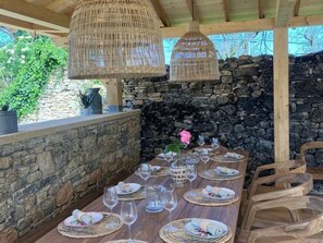 A wooden dining table set with plates, glasses, and napkins, under woven pendant lights, with a stone wall and greenery in the background. - Peaceful house with private pool and pets allowed (Florimont-Gaumier)