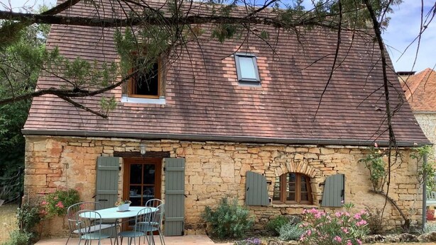 Une maison en pierre avec un patio carrelé, une table entourée de chaises et un petit jardin agrémenté de fleurs roses.