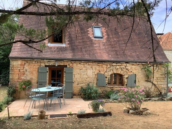A stone house with a tiled patio, a table with chairs, and a small garden with pink flowers.