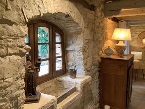 A rustic interior with a stone wall, a wooden arched window, a small table with a lamp, and a statue on the windowsill. - Peaceful house with private pool and pets allowed (Florimont-Gaumier)