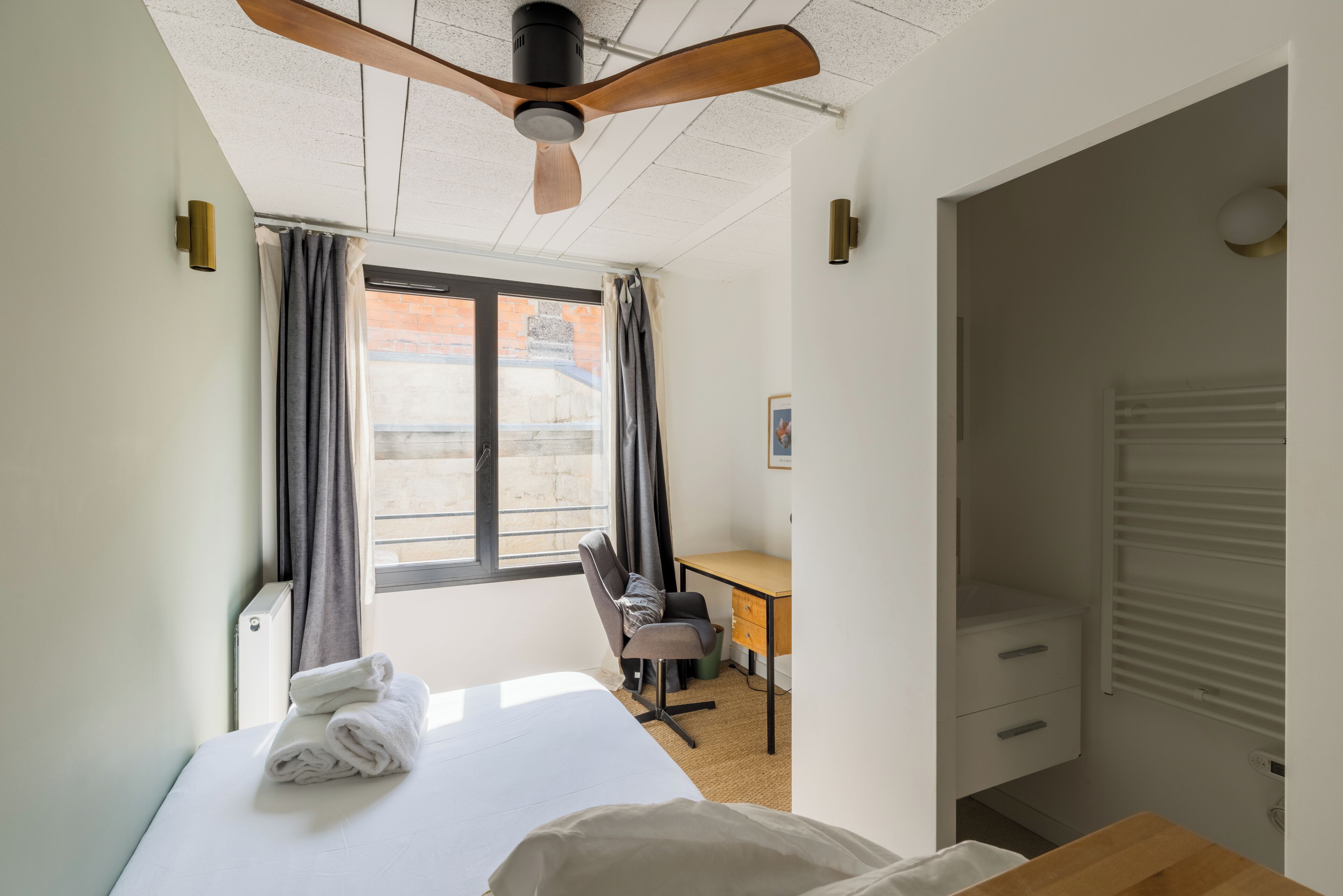 Une chambre d’hôtel avec un lit, un bureau et une chaise. Il y a une fenêtre avec des rideaux, un ventilateur de plafond et un tableau au mur.