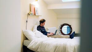 A person using a laptop on a bed in a room with a skylight, a round window, and a radiator. - Outsite Bordeaux - Work, Connect, Explore (Bordeaux)