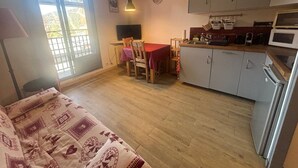A compact living space with a kitchenette, a dining area with a red table and chairs, a bed with patterned bedding, and a window with a view of greenery.