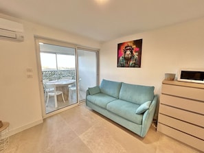 Uma sala de estar com um sofá azul claro, uma televisão sobre um móvel de madeira e uma porta de vidro deslizante que dá para uma área externa com mesa e cadeiras.