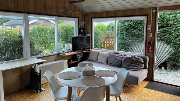 Um quarto aconchegante com teto de madeira, sofá, mesa de jantar com cadeiras, TV e janelas amplas com vista para o verde lá fora.