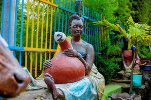A sculpture of a woman holding a large pot, set against a colorful fence and greenery. - Kivu Nest Hotel  (Gisenyi)