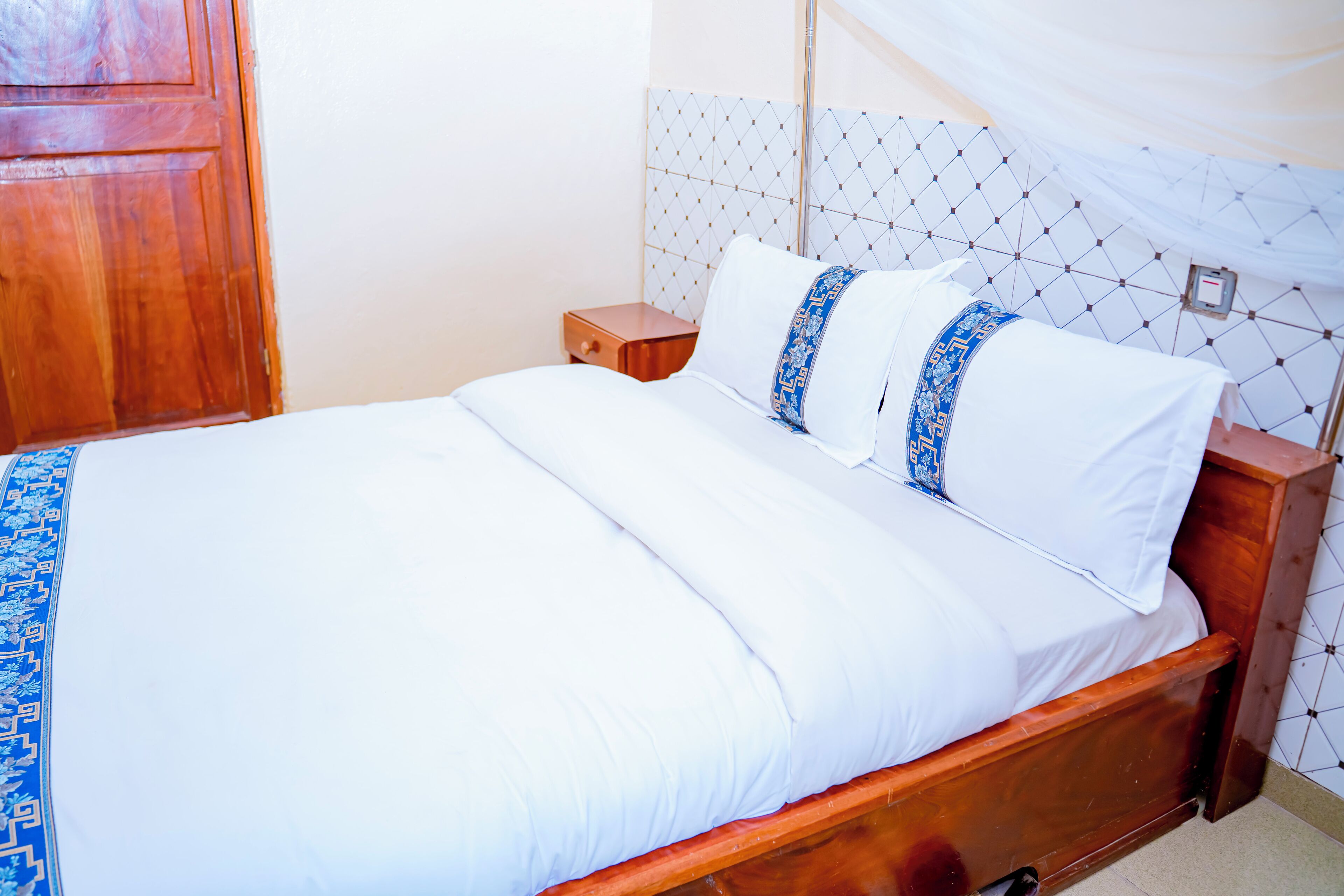 Un letto rifatto con lenzuola bianche e cuscini decorativi blu, un comodino in legno e una parete piastrellata con motivo geometrico.