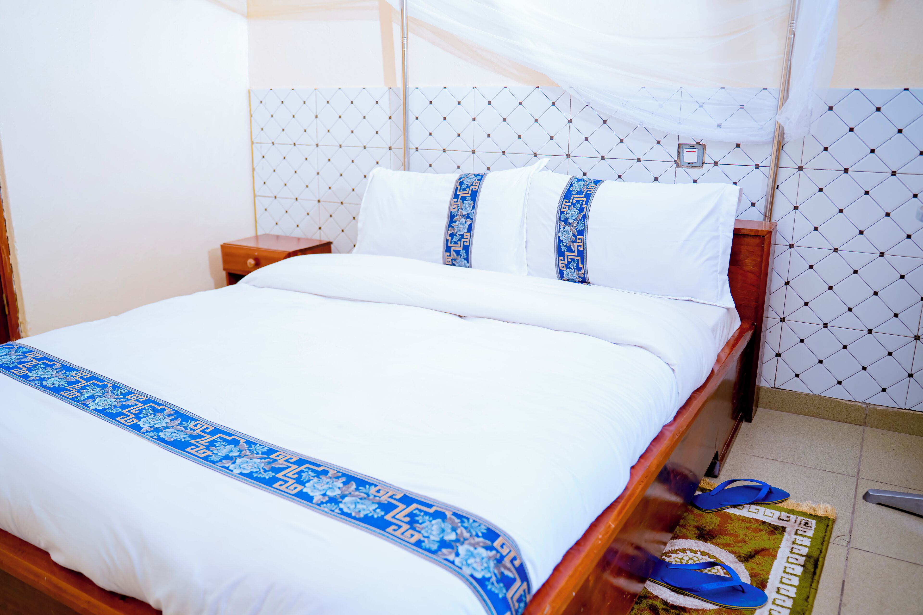 Una cama bien hecha con una colcha blanca y azul, un cabecero de madera y una mesita de noche con una lámpara.