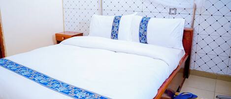 Una cama bien hecha con una colcha blanca y azul, un cabecero de madera y una mesita de noche con una lámpara.