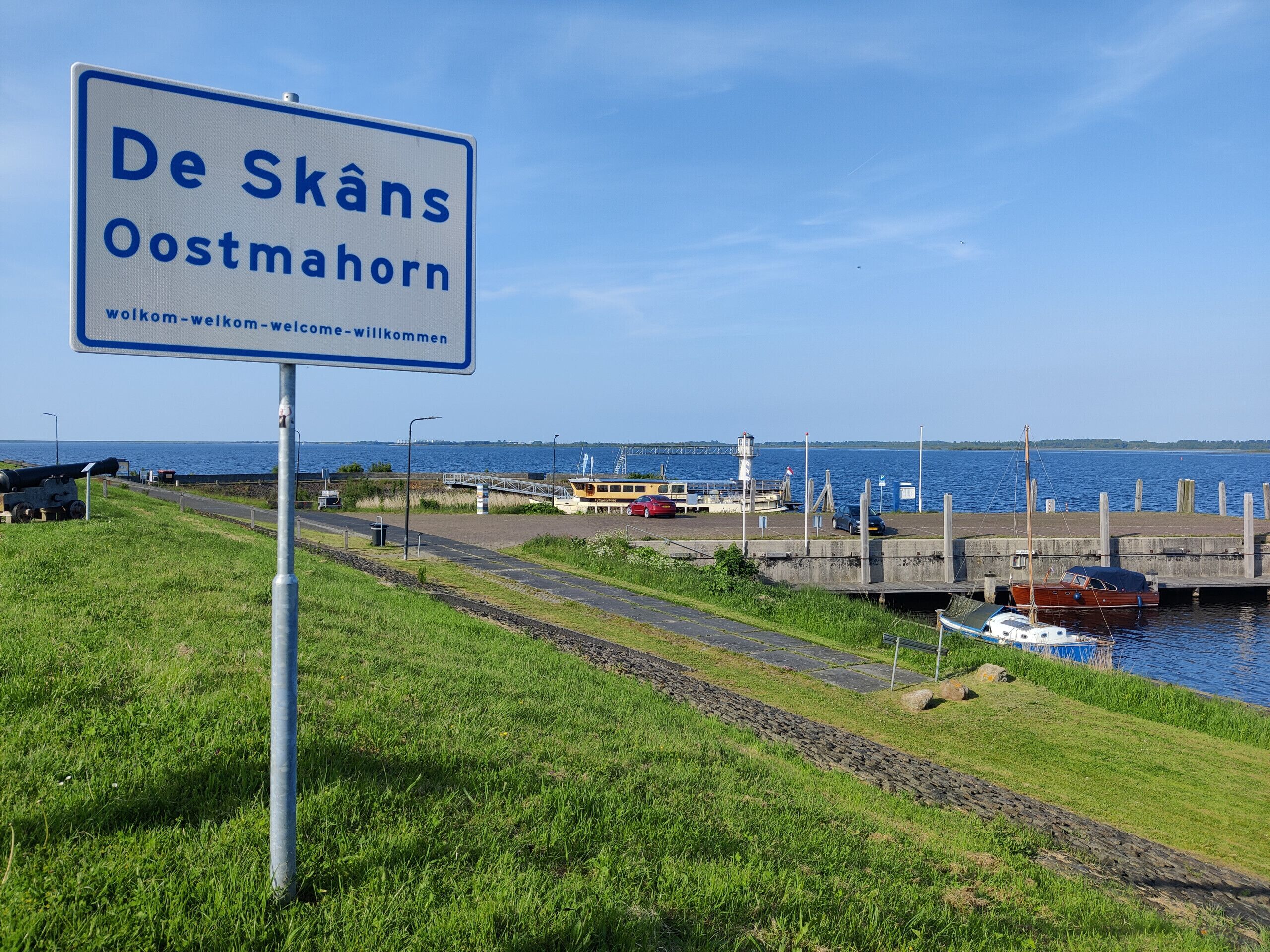 Une pancarte indiquant « De Skåns Oostmohorn » avec un message d’accueil, une zone herbeuse, une route, une étendue d’eau et des bateaux amarrés à un quai.