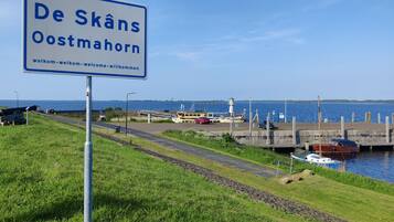 Une pancarte indiquant « De Skåns Oostmohorn » avec un message d’accueil, une zone herbeuse, une route, une étendue d’eau et des bateaux amarrés à un quai.