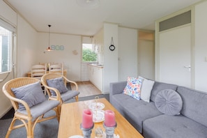 A cozy living area with a grey sofa, wicker chairs, a wooden table with candles, and a kitchen in the background.
