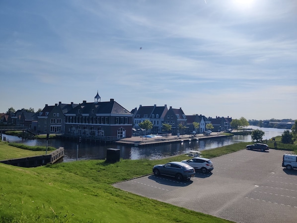 A canal-side building with a waterway, parked cars, and a grassy area.