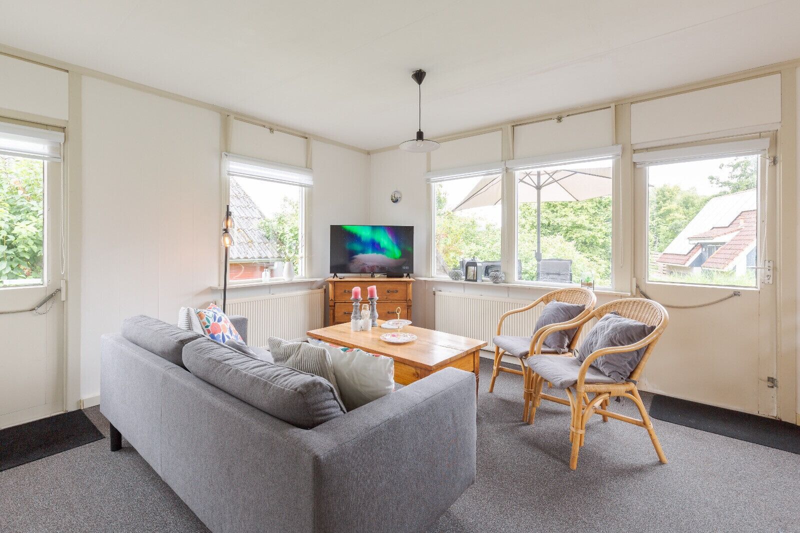 Un salón con un sofá, dos sillones de mimbre, una mesa de madera, un televisor y ventanas grandes.