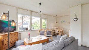 A cozy living room with a sofa, wooden coffee table, and a TV. There are windows, a dining area, and a ceiling fan.