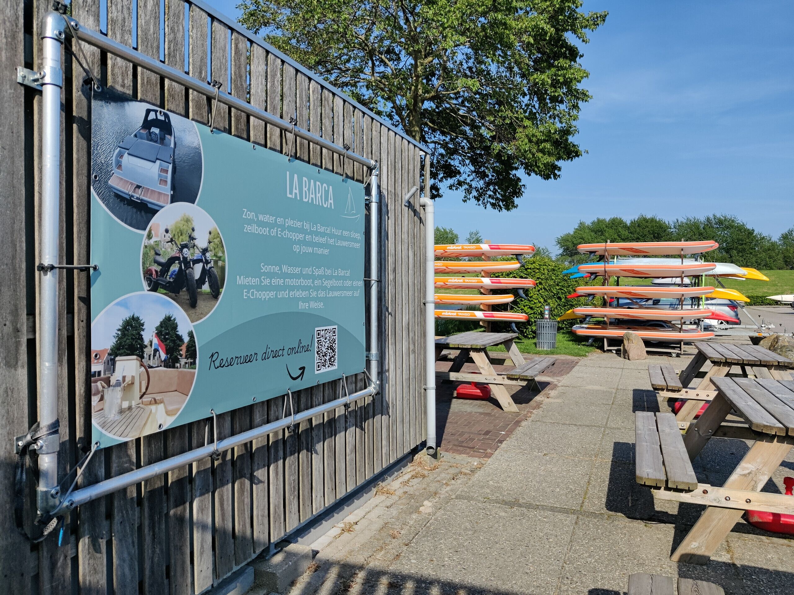 A wooden fence with a signboard advertising La Barca, featuring a QR code and images of a motorboat, a motorcycle, and a scenic view.