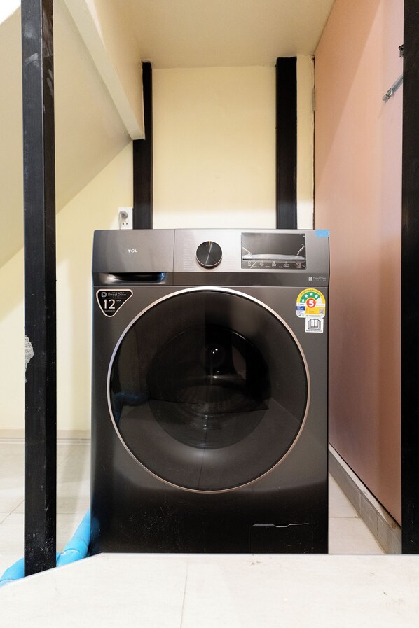 A front-loading washing machine in a narrow hallway with a tiled floor and a door on the right.