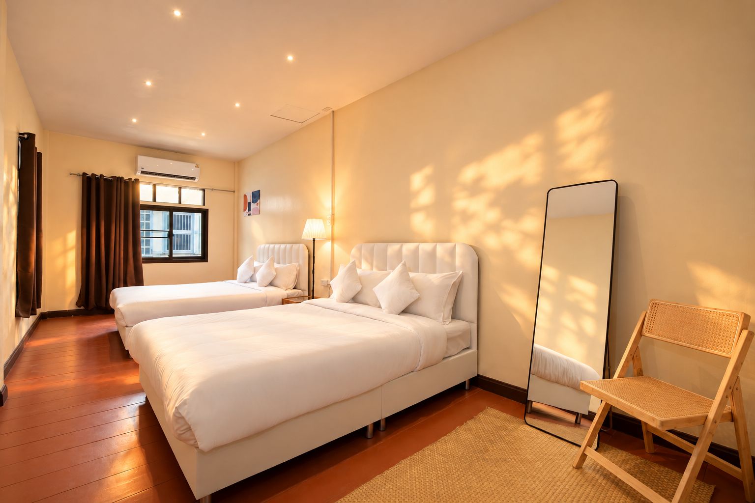 Habitación de hotel con dos camas, un espejo de pie, una silla de madera y una ventana con cortinas.