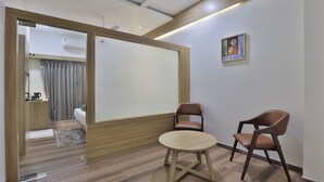 A modern room with a wooden floor, a round table, two brown chairs, a white sofa, and a sliding door leading to a bathroom. - Hotel SR Laxmi (Anand)