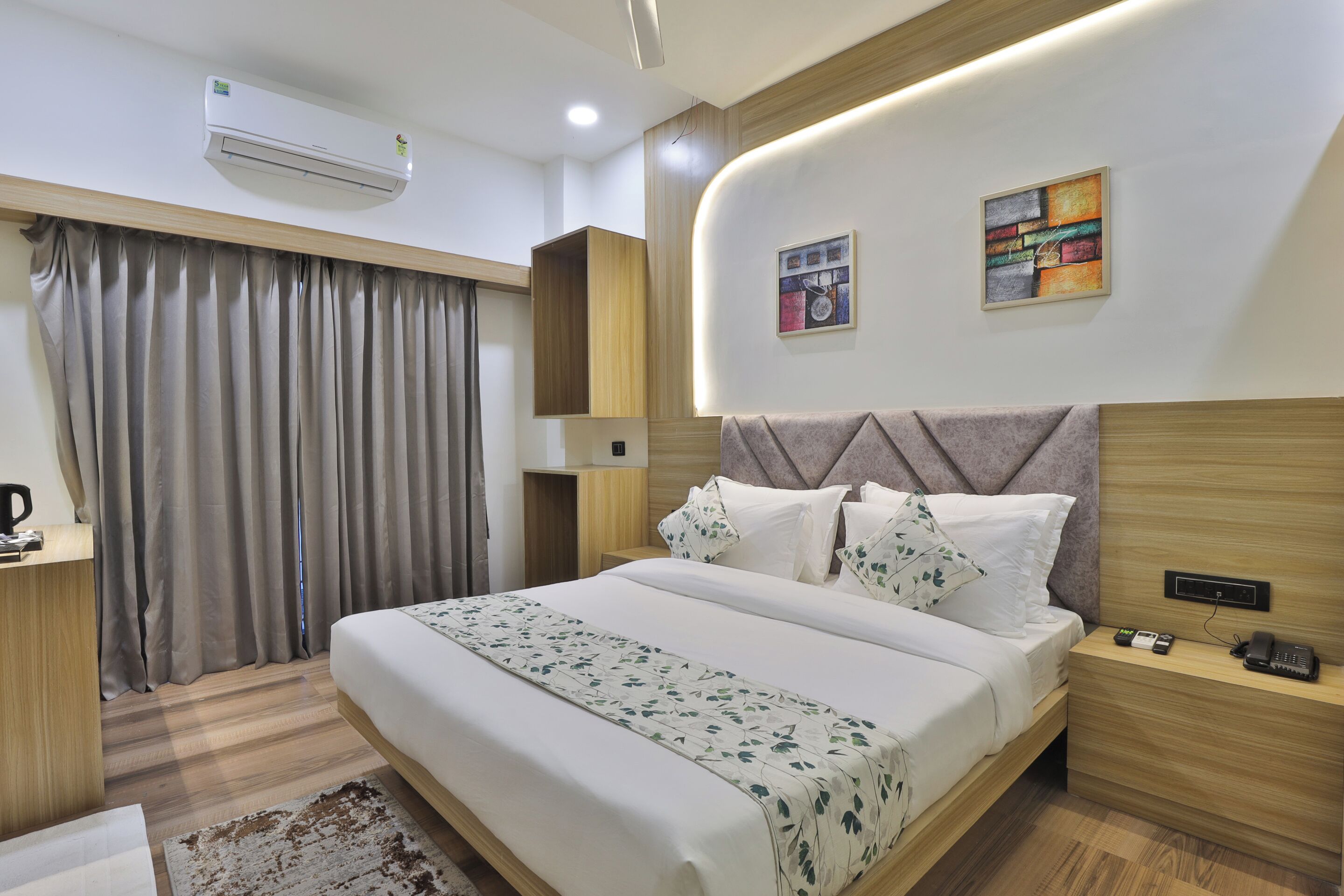 A modern hotel room with a large bed, bedside tables, an air conditioning unit, and a painting on the wall.