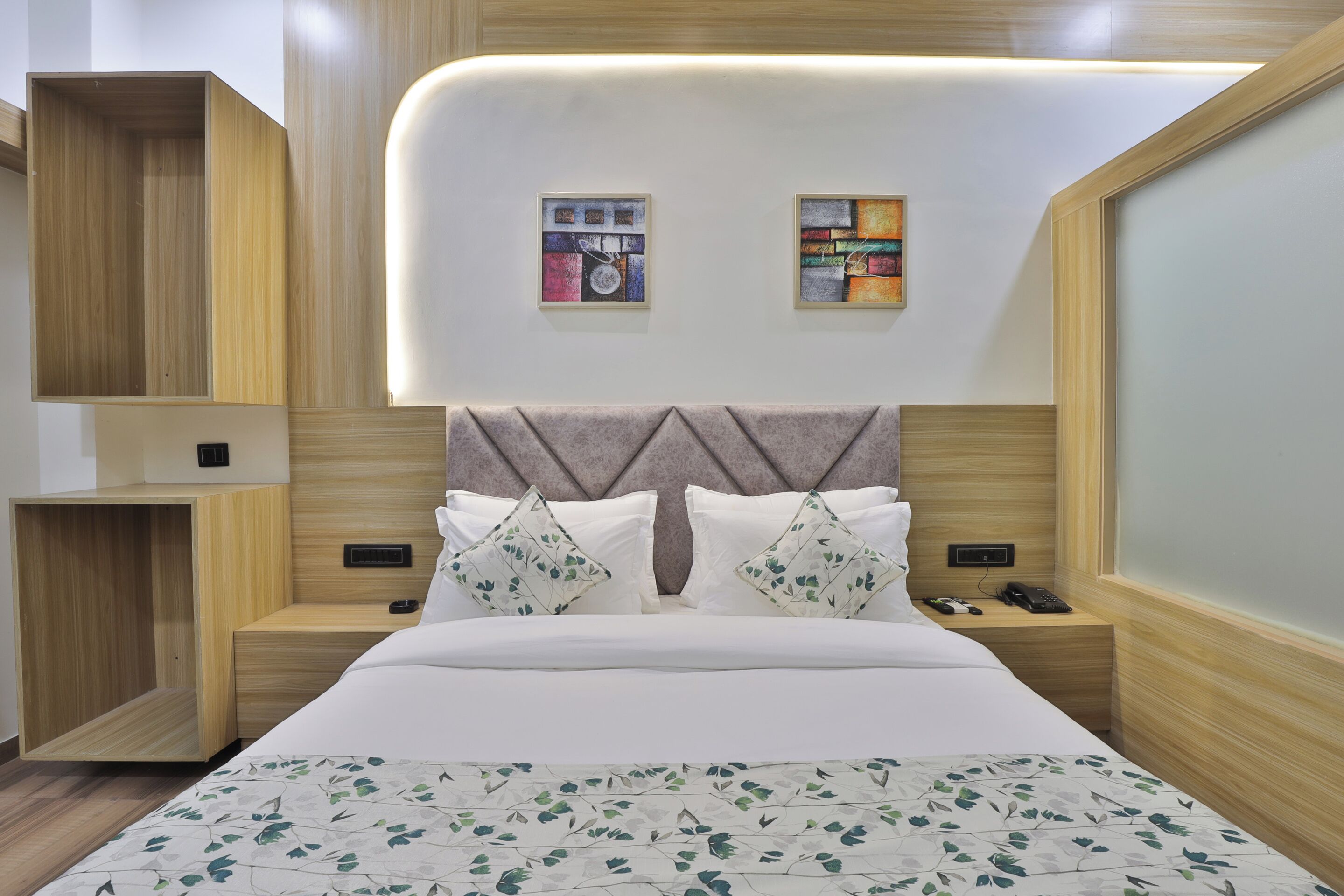 A modern hotel room with a wooden headboard, a bed with white bedding, two bedside tables, and two wall-mounted artworks.