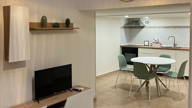 A modern living room with a flat-screen TV, a sofa, a dining area with a round table and chairs, and a kitchen in the background. - Cavour House Lentini (Lentini)