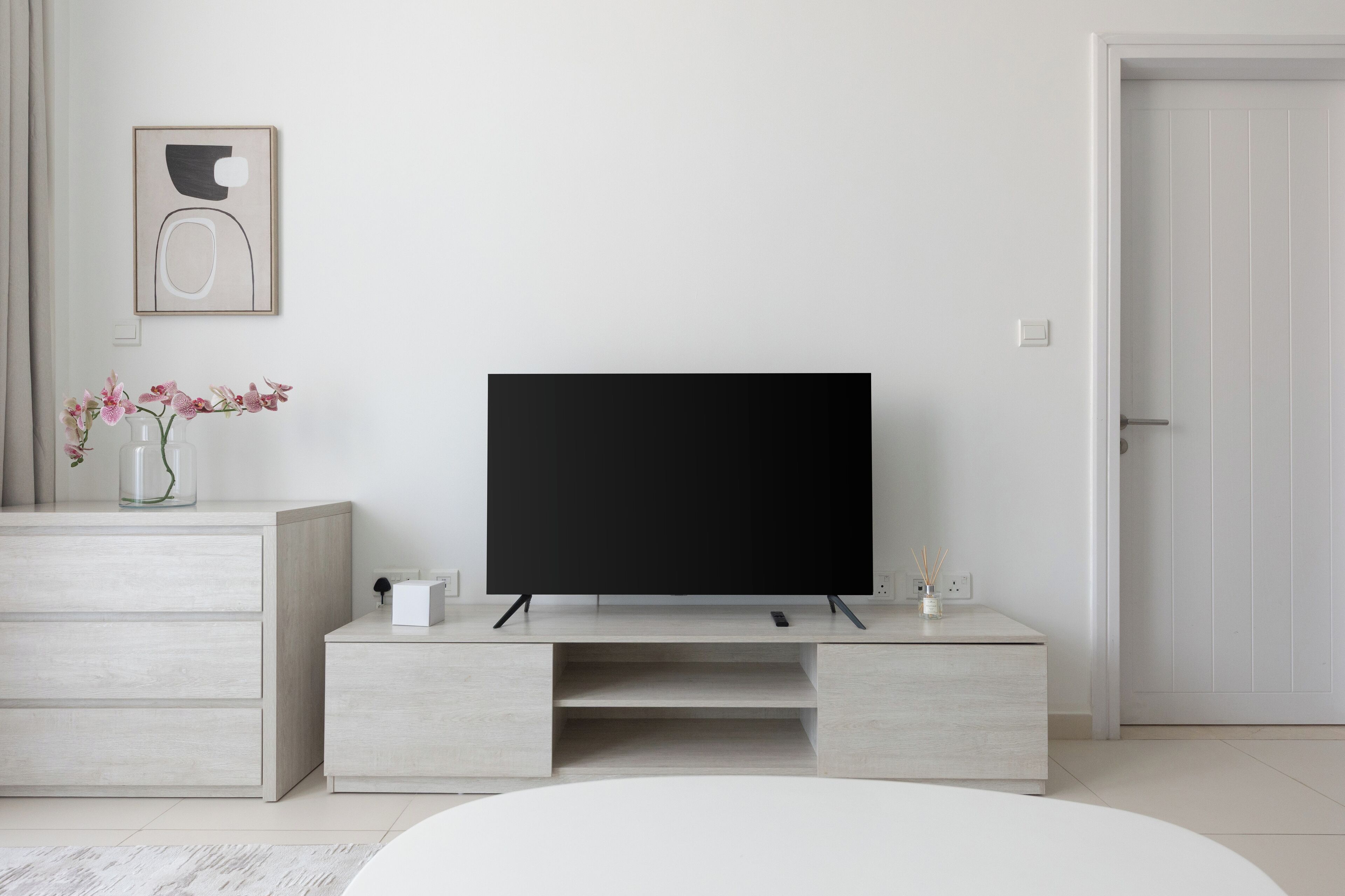 A modern living room with a flat-screen TV on a wooden stand, a vase with flowers, and a white door.