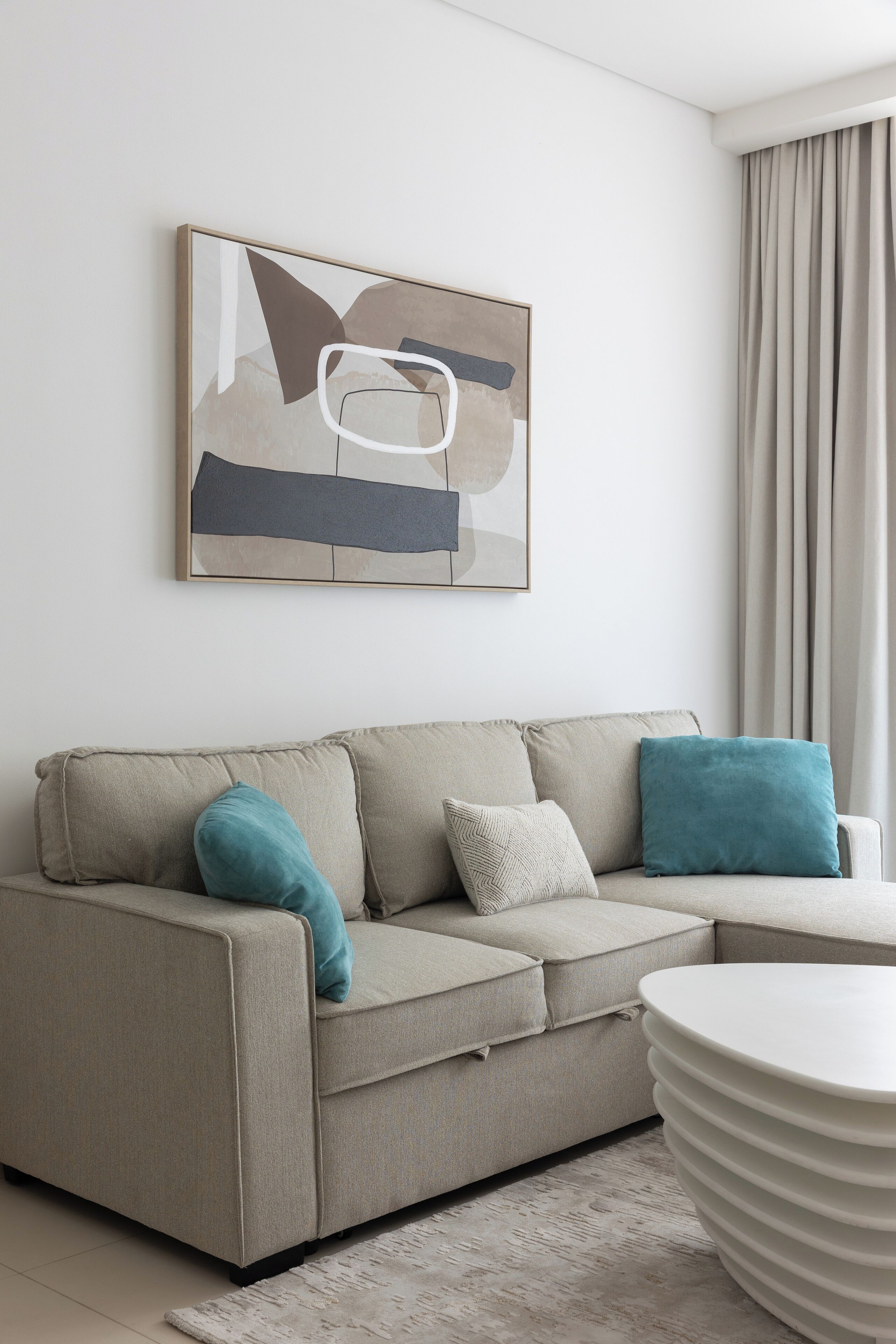 Ein modernes Wohnzimmer mit einem beigefarbenen Sofa, Kissen in Türkis und Weiß, einem weißen Couchtisch und einem modernen, abstrakten Gemälde an der Wand.