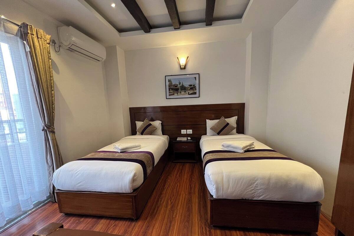 Habitació d'hotel amb dos llits, sòl de fusta, sostre amb bigues descobertes, un aparell d'aire condicionat i un quadre encimbellat a la paret.