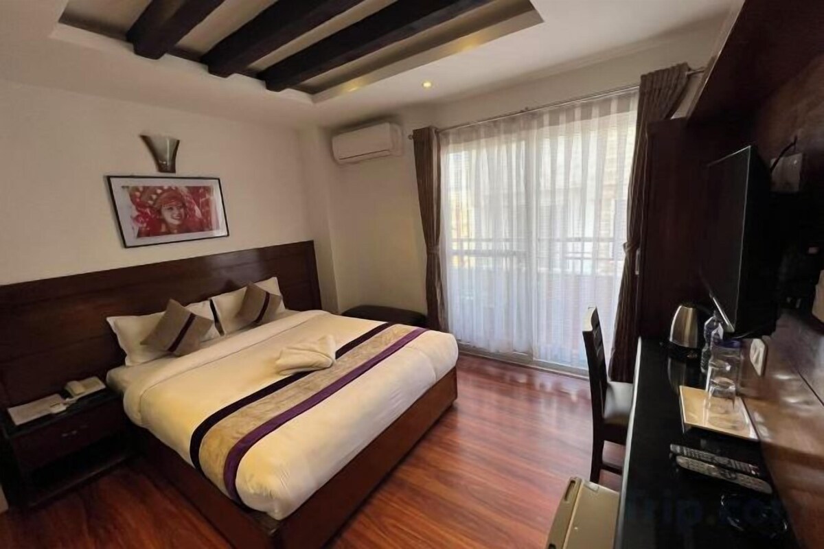 A hotel room with a large bed, a wooden headboard, a bedside table with a lamp, a flat-screen TV, and a window with curtains.