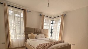 A bedroom with a large bed, beige curtains, and a green patterned rug. - Lux Suites The comfort Place Naivasha (Naivasha)