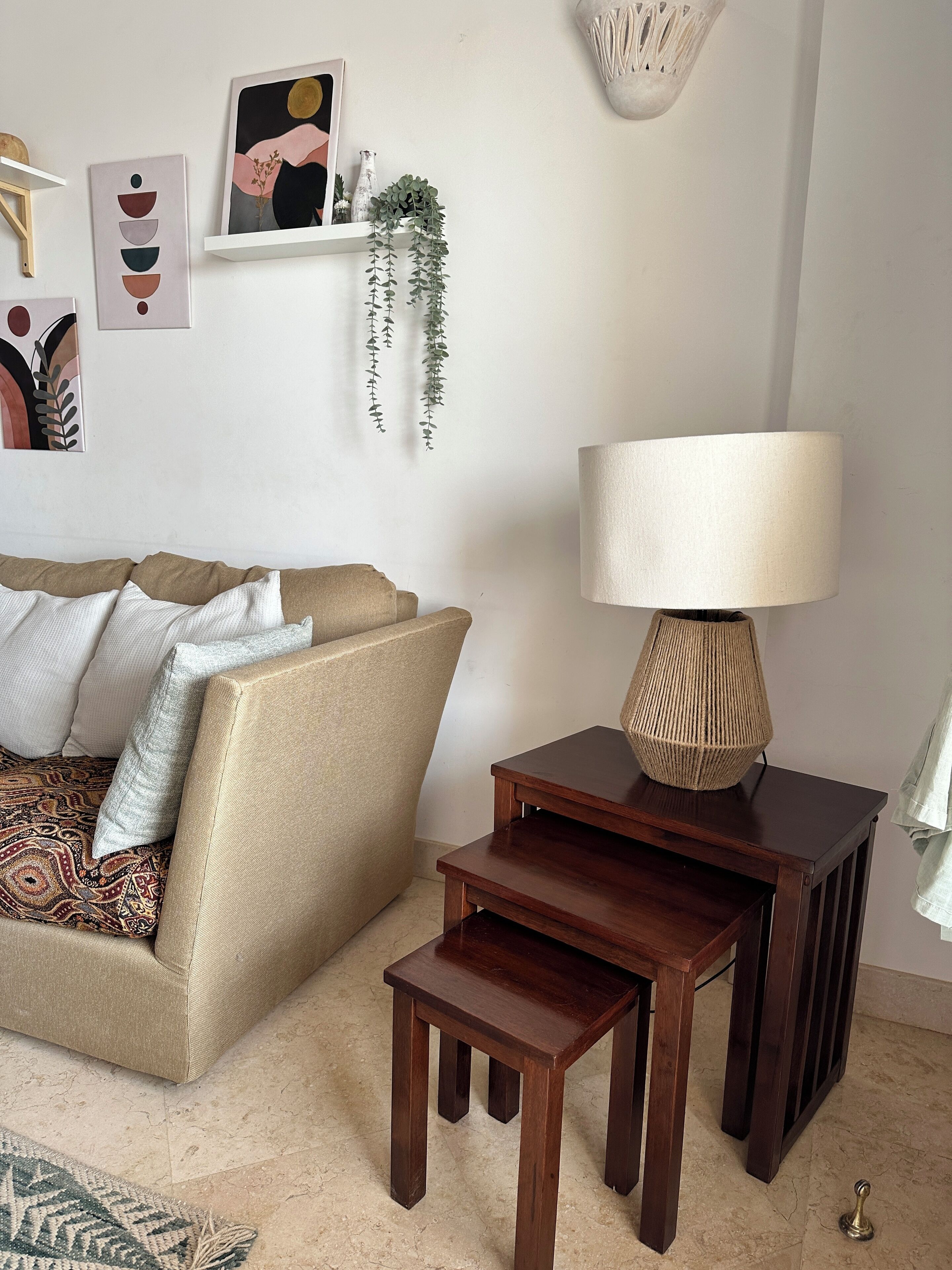 A living room with a beige sofa, patterned cushions, a wooden side table with a lamp, and a small wooden stool.