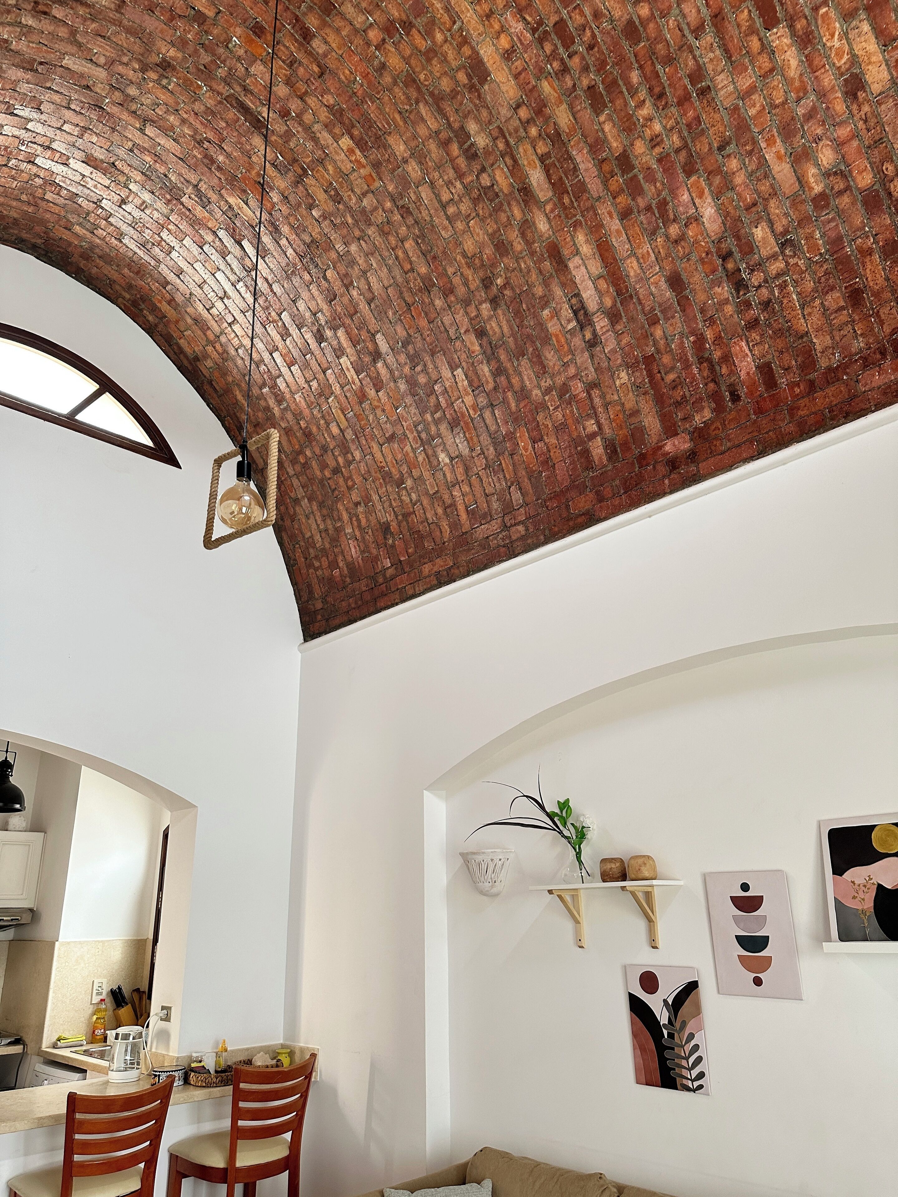 A room with a brick ceiling, white walls, and a kitchen area. There are two wooden chairs, a white shelf with decorative items, and a framed picture on the wall.
