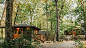 A rustic cabin with a wooden deck, surrounded by tall trees and a gravel path. - Harriman Outdoor Center - Cranberry Cabin (Haverstraw)