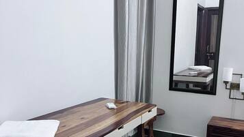 A room with a wooden massage table, a small bedside table, a mirror, and a curtain.