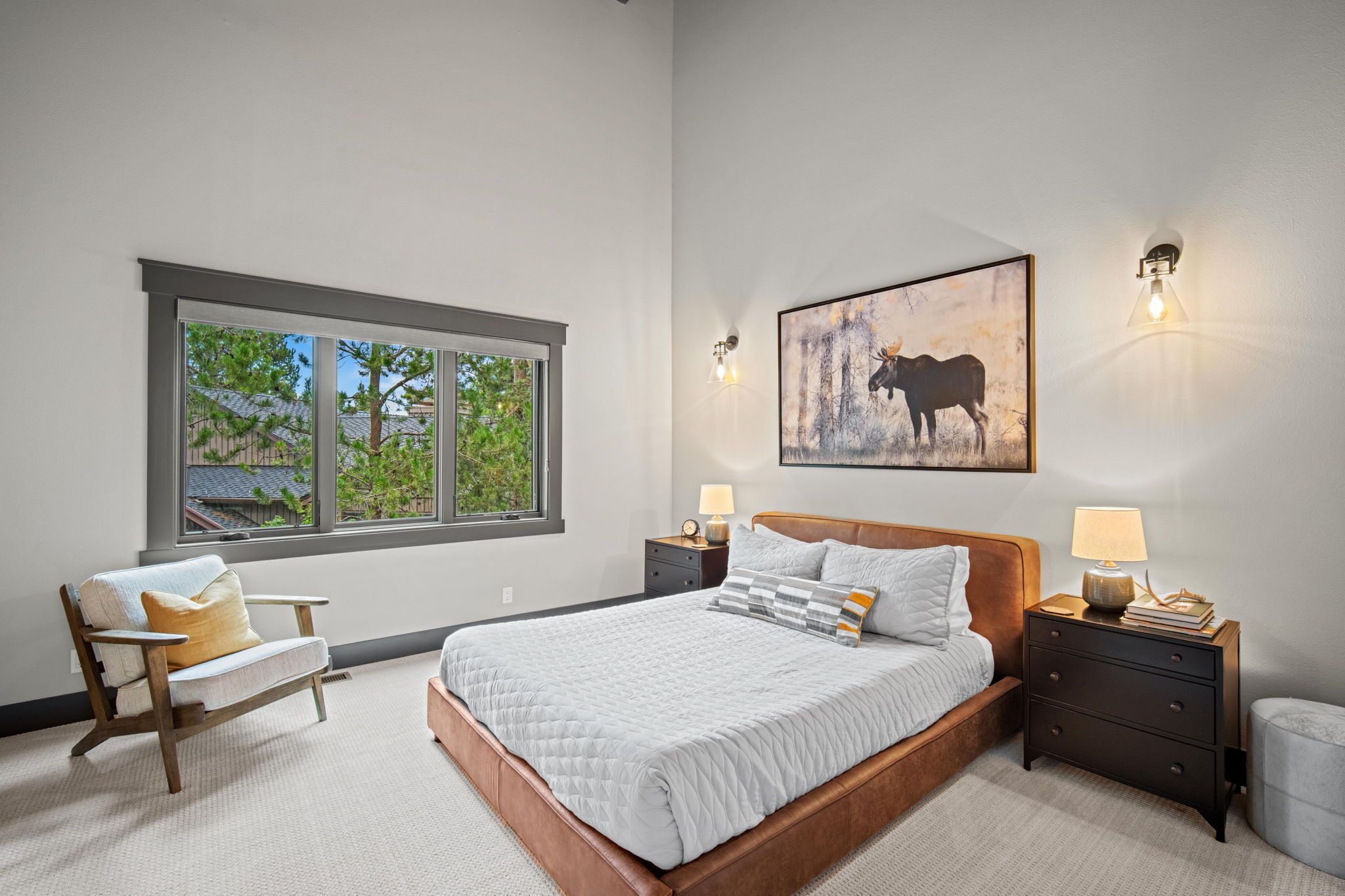 Une chambre à coucher avec un grand lit, une table de chevet avec une lampe, une chaise et une fenêtre donnant sur des arbres.