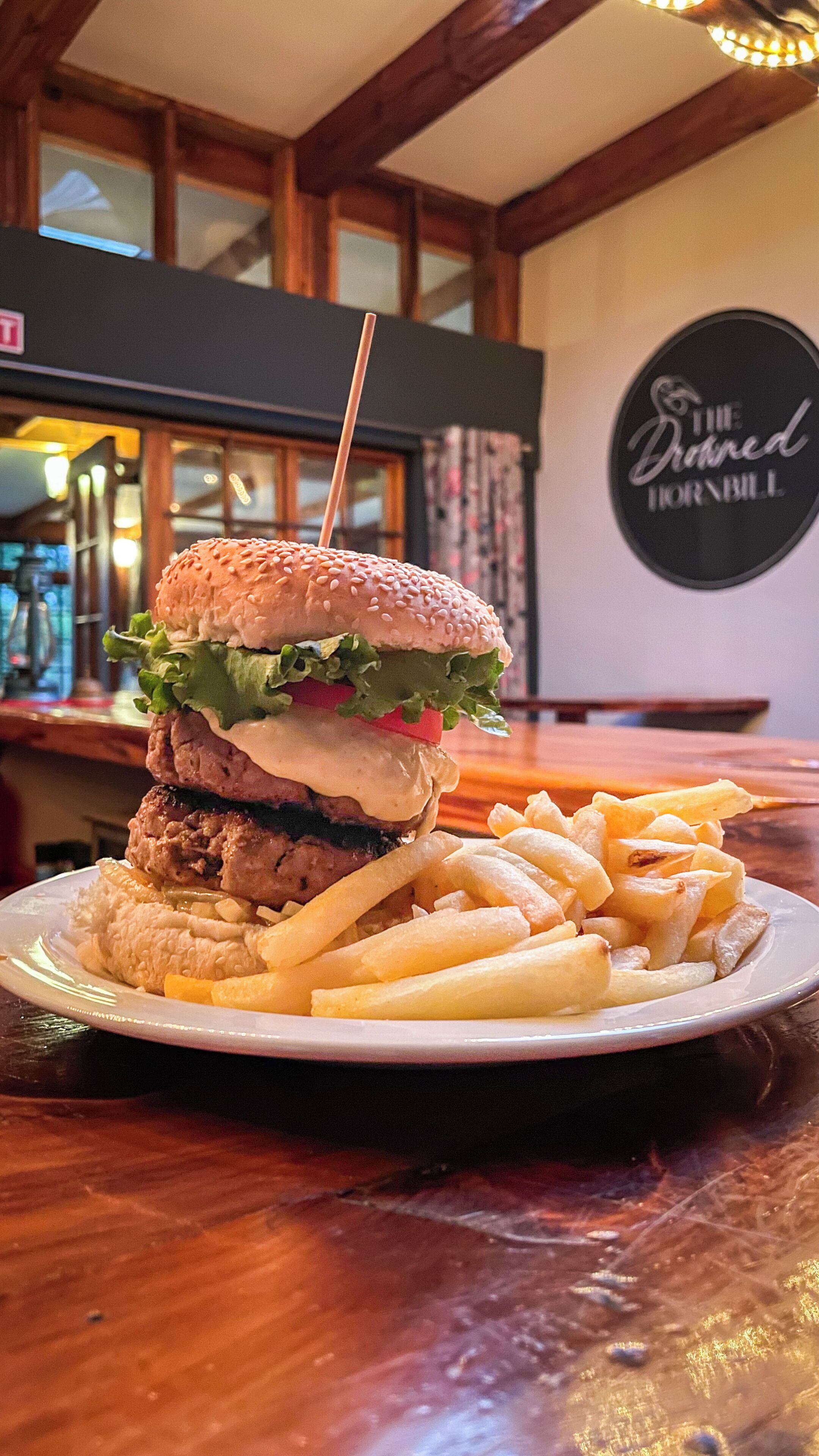 A burger with fries on a plate, a wooden table, and a wall with a sign reading "The Drowned Hornbill".