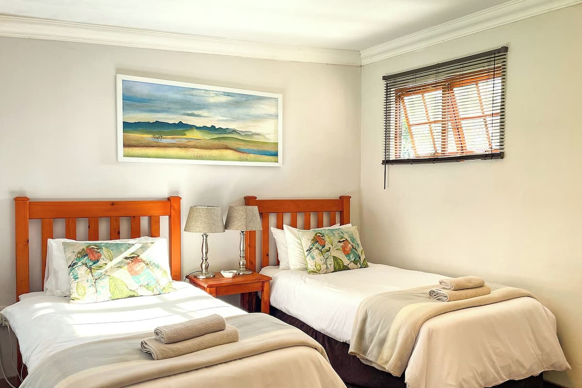 A bedroom with two beds, a painting, and a window with blinds.