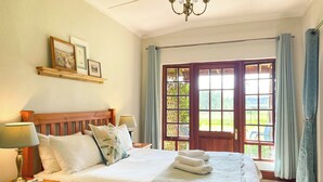 A well-lit bedroom with a wooden bed, a nightstand with a lamp, a window with curtains, and a view of greenery outside. - Molweni Kamberg Valley Bed and Breakfast (Elandskop)