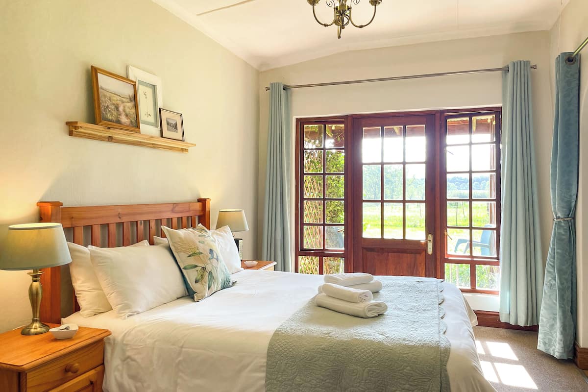 A well-lit bedroom with a wooden bed, a nightstand with a lamp, a window with curtains, and a view of greenery outside.