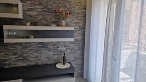 Una sala moderna con pared estampada en piedra, repisa flotante con adornos, consola oscura y ventana con cortinas traslúcidas.