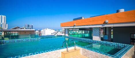 A rooftop infinity pool with a wooden deck and a stone wall, overlooking a cityscape.
