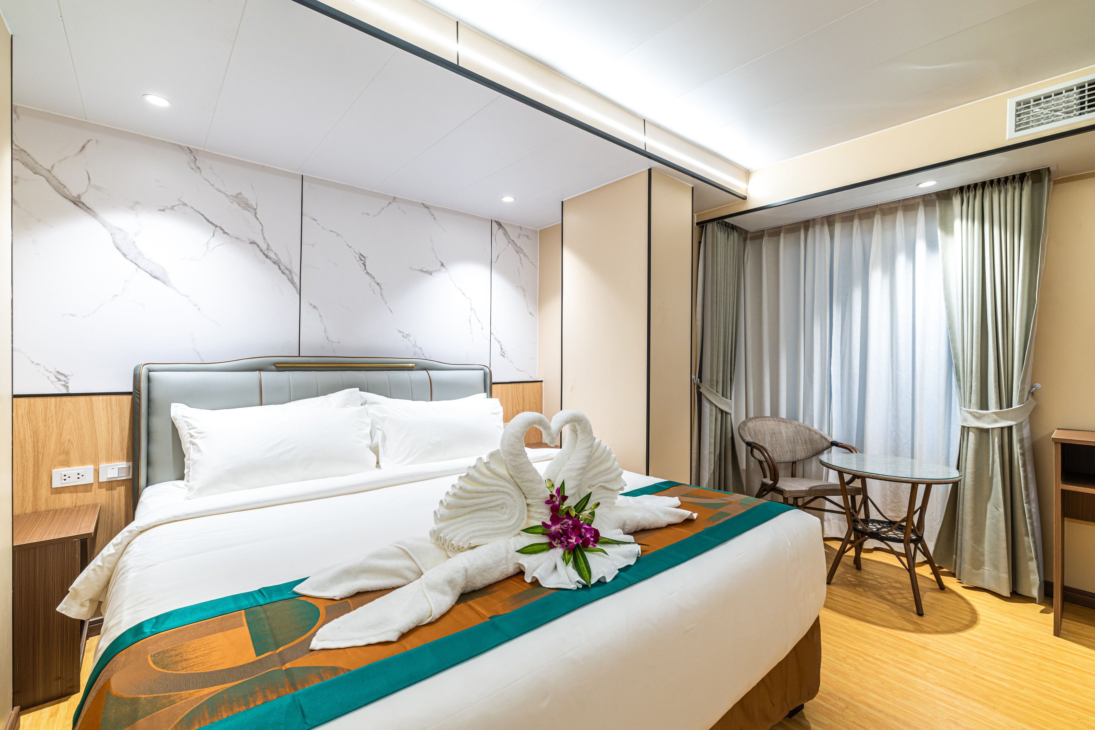 A hotel room with a neatly made bed, a desk with a chair, and a view of the outside through curtains.