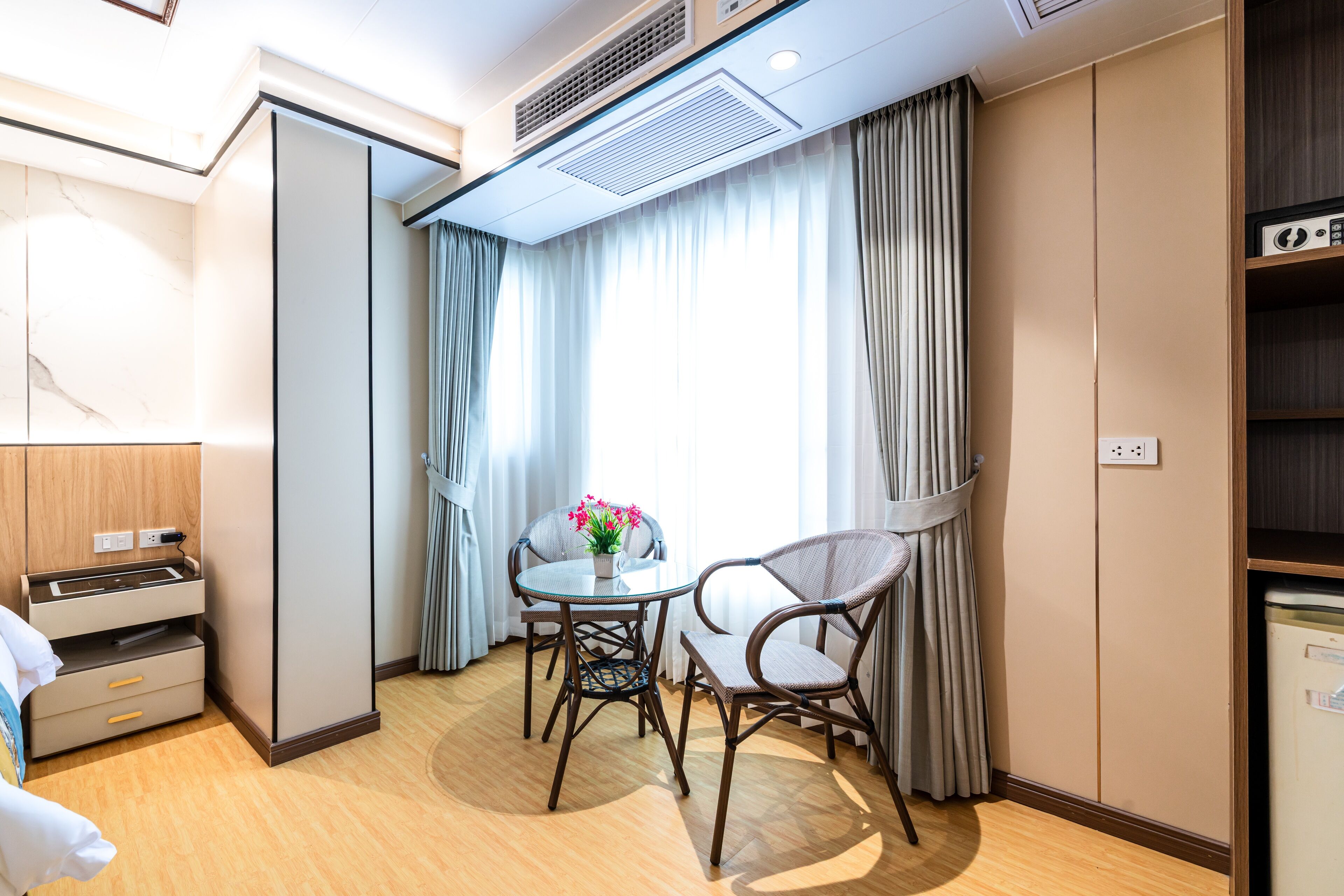 Habitación de hotel pequeña y funcional con cama, una mesita con sillas, una planta en maceta y ventana con cortinas.