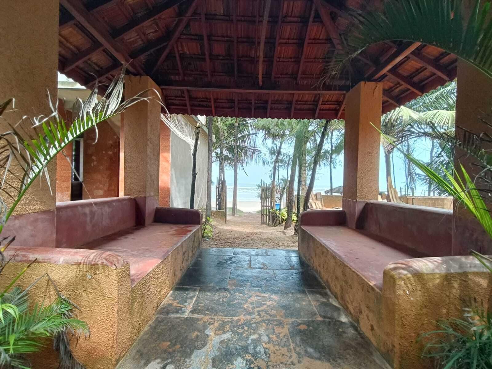 A covered outdoor seating area with stone flooring and wooden benches, surrounded by palm trees and a view of the beach.